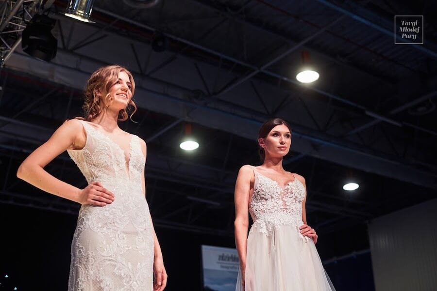 Dwie modelki w białych sukniach ślubnych prezentują kreacje na wybiegu podczas targow slubnych.